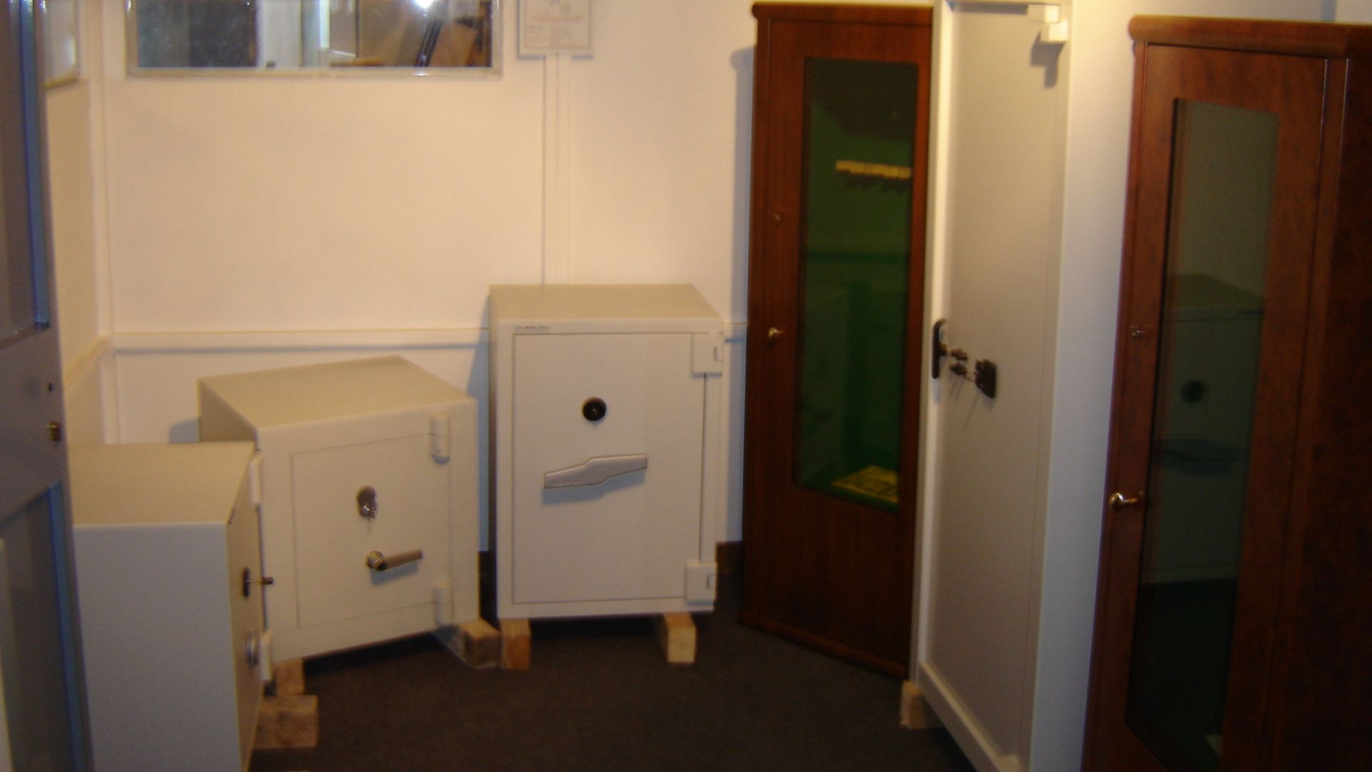 Dans une pièce aux murs blancs, quatre coffres-forts de tailles différentes, dont deux posés sur des blocs de bois. Une armoire en bois brun foncé à l'intérieur vert.