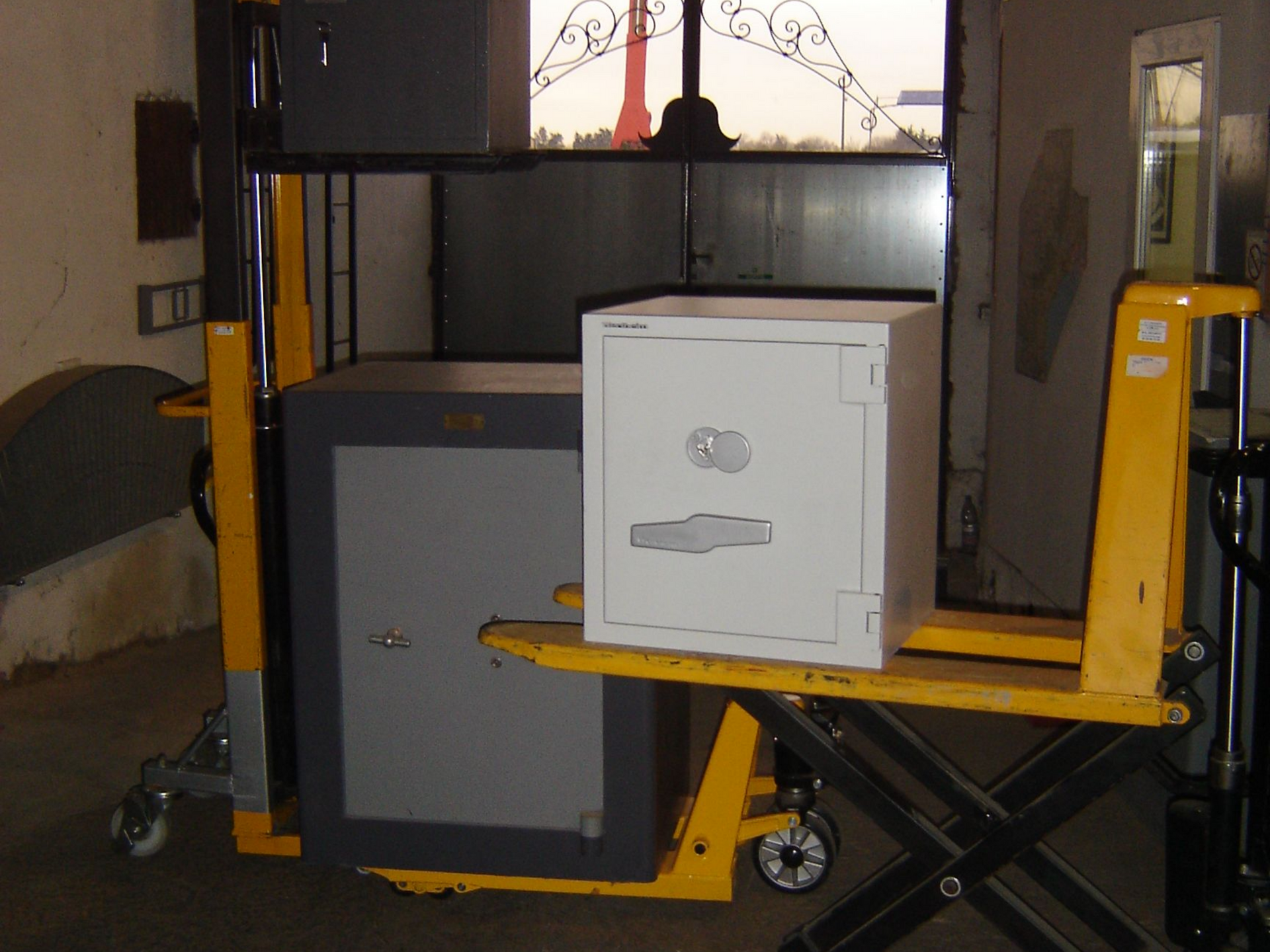 Un coffre-fort est posé sur un transpalette jaune. Un autre coffre-fort se trouve à proximité, dans un environnement industriel.