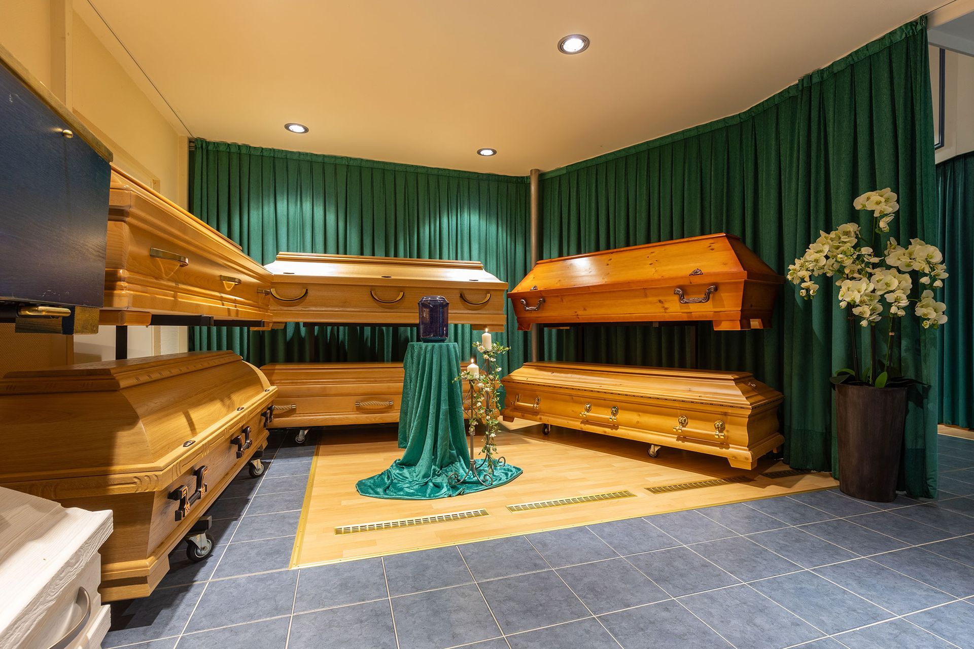 Holzsärge, ausgestellt in einem Ausstellungsraum mit grünen Vorhängen und einem Blumenarrangement.