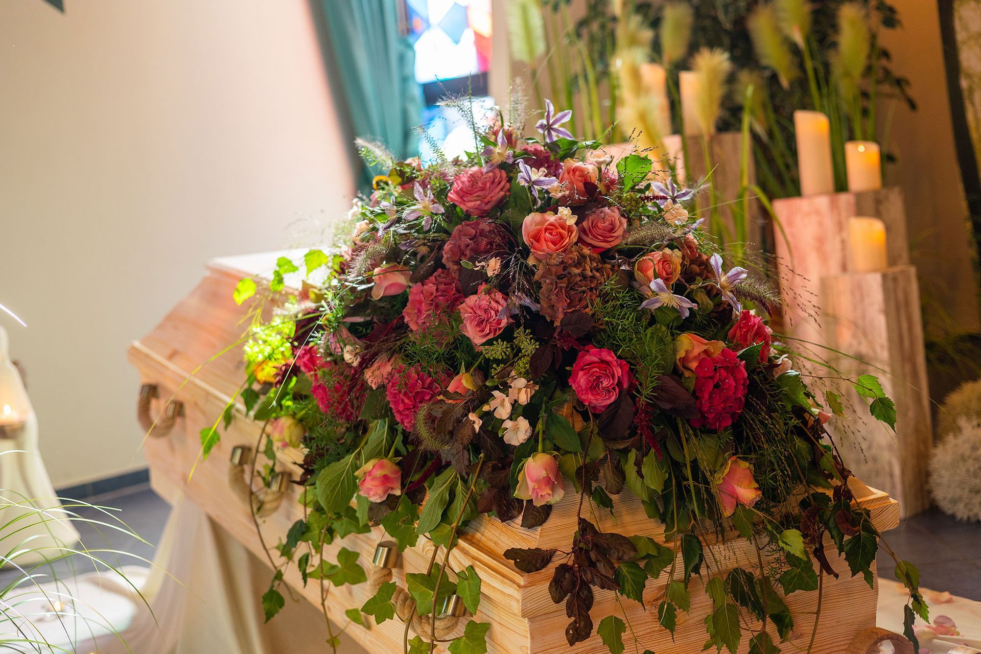 Ein mit roten und rosa Blumen geschmückter Holzsarg. Innenaufnahme, im Hintergrund Kerzen.