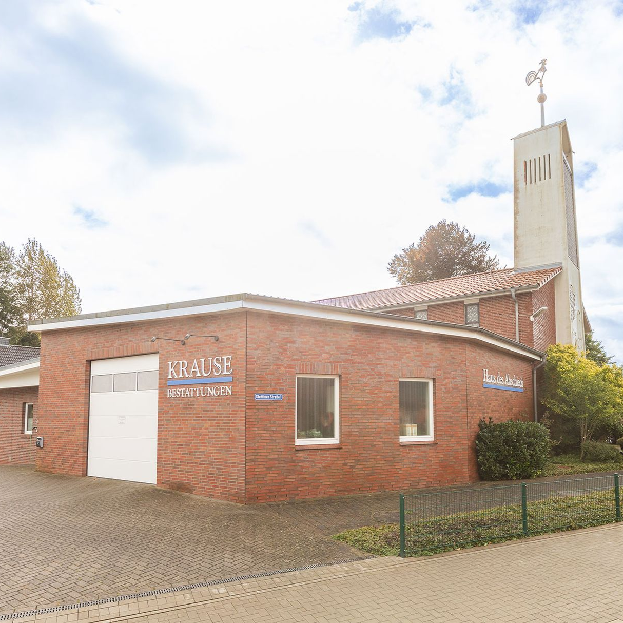 Backsteingebäude mit weißem Garagentor und hohem, weißem Turm; darüber bewölkter Himmel.