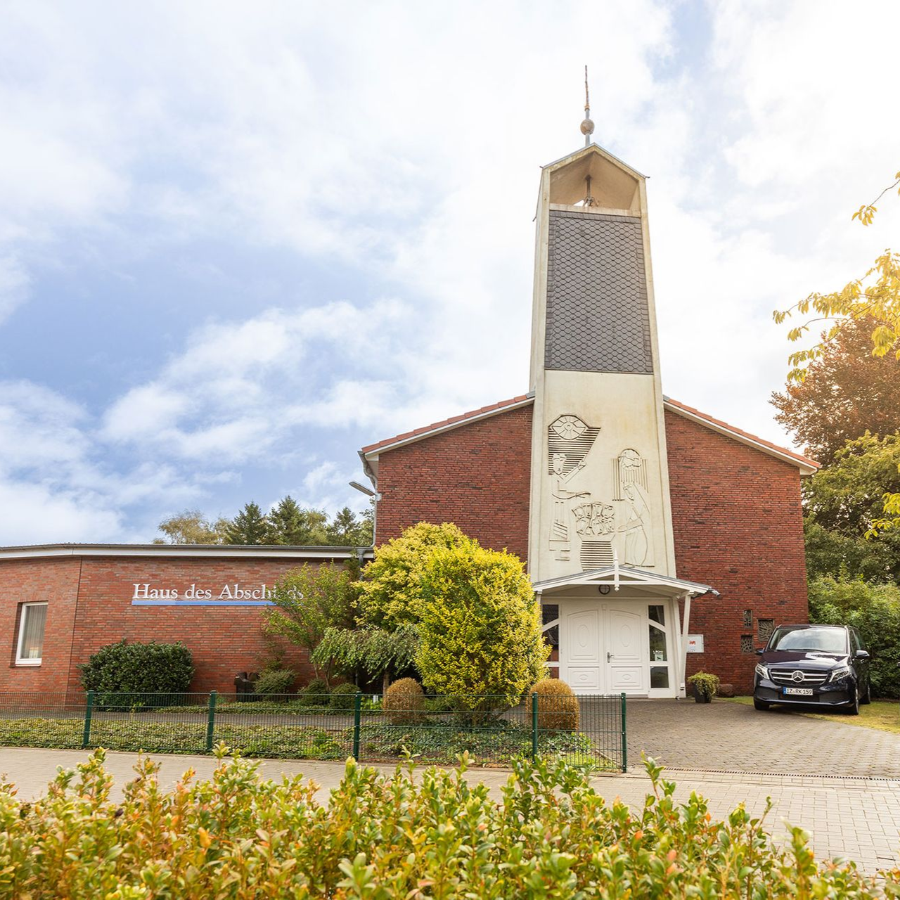 Kirche mit hohem, weißem Glockenturm und Backsteingebäude. Vor dem Gebäude ist ein Auto geparkt.