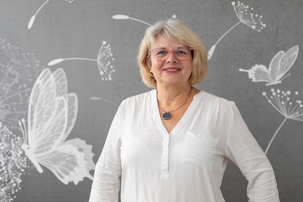 Frau in weißem Hemd lächelt, steht vor einer grauen Wand mit Schmetterlingszeichnungen.