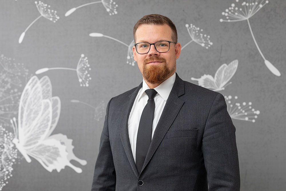 Ein Mann im Anzug und mit Brille steht vor einer grauen Wand mit weißen Schmetterlings- und Löwenzahnmotiven.