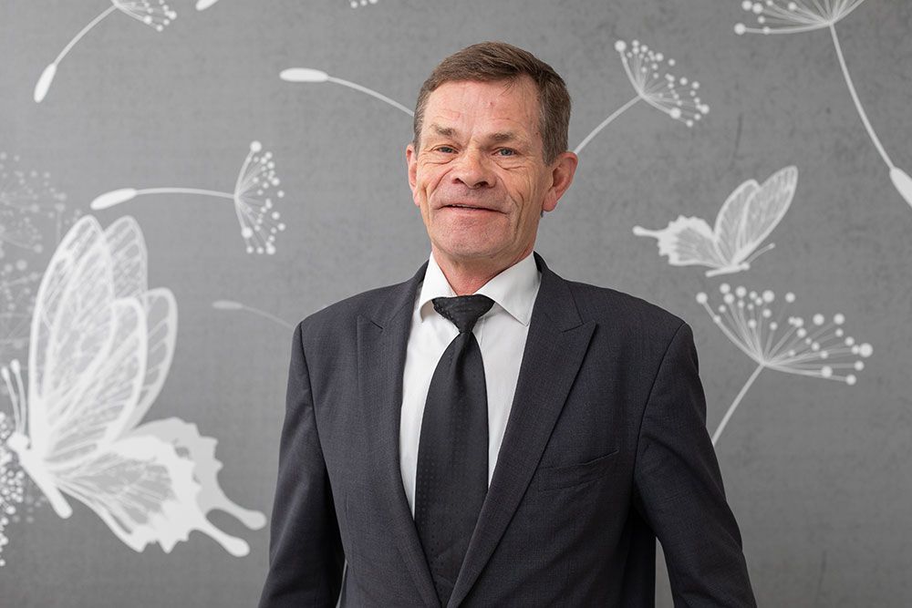 Ein Mann im Anzug steht vor einer grauen Wand mit weißen Schmetterlingsmotiven.