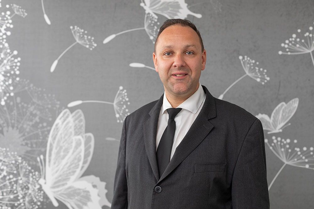 Ein Mann im Anzug lächelt vor einer grauen Wand mit weißen Schmetterlings- und Löwenzahnmotiven.