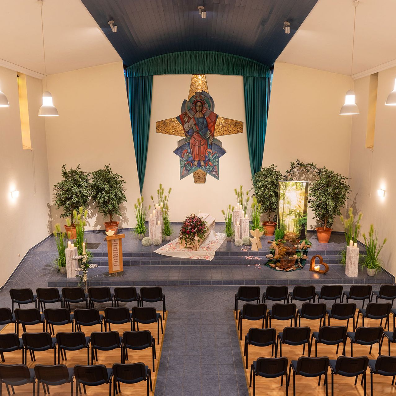 Innenansicht einer Kapelle mit Stuhlreihen, einem Blumenarrangement und einem Kreuz an der Wand.