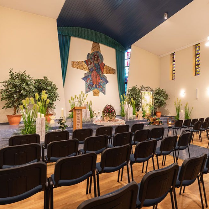 Innenraum eines Kirchenraums mit Stühlen, Altar, Kunstwerken und Pflanzen. Blaue, grüne und neutrale Farbtöne.