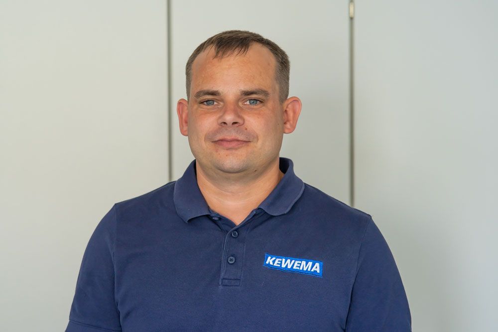 Ein Mann in einem blauen Hemd mit der Aufschrift „Kewema“ steht vor einer weißen Wand.