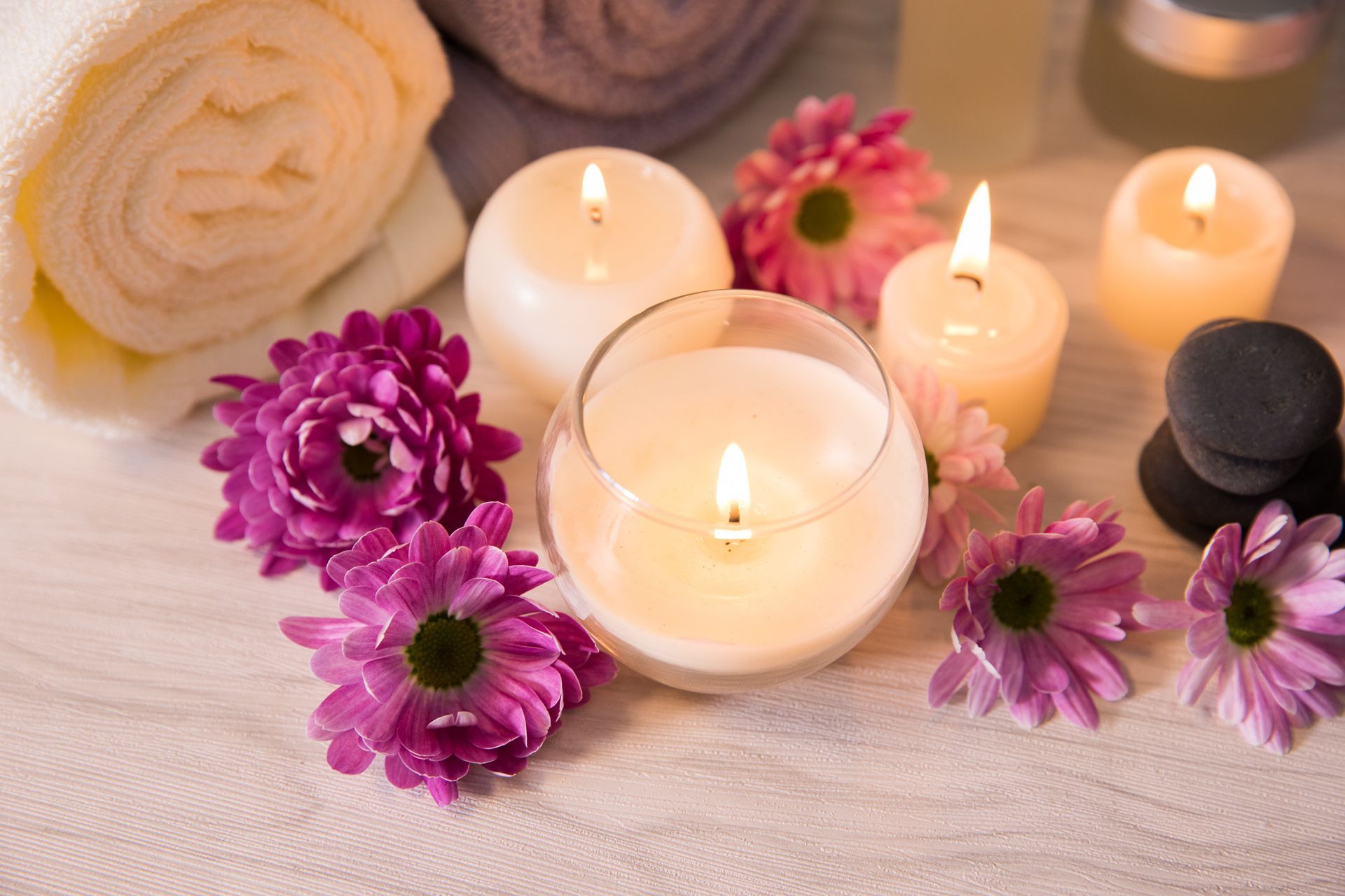 Scenografia spa: płonące świece, kwiaty, zwinięte ręczniki i czarne kamienie na teksturowanej białej powierzchni.