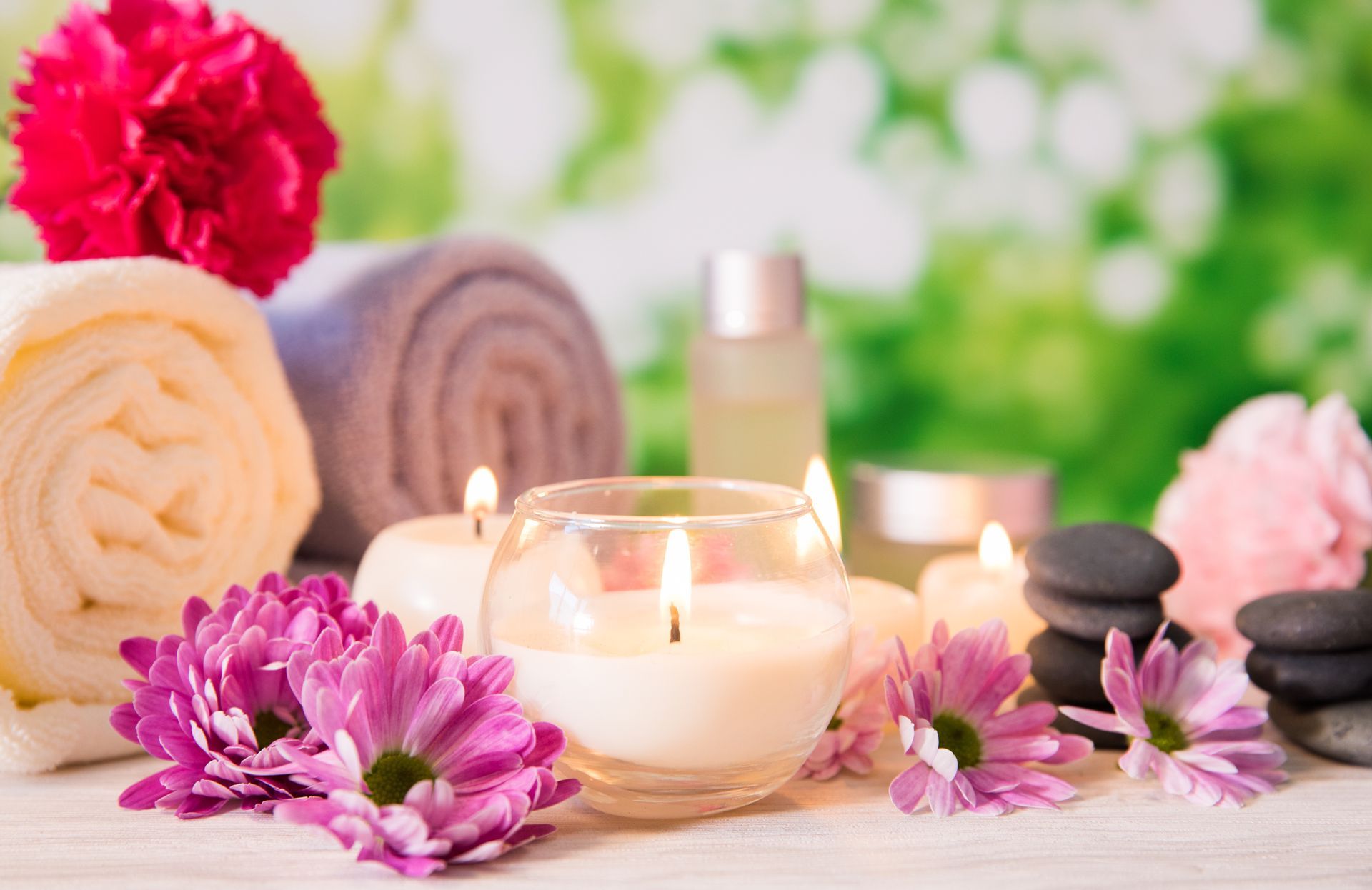 Aranżacja spa ze świecami, zwiniętymi ręcznikami, kwiatami i kamieniami na drewnianej powierzchni z rozmytym zielonym tłem.