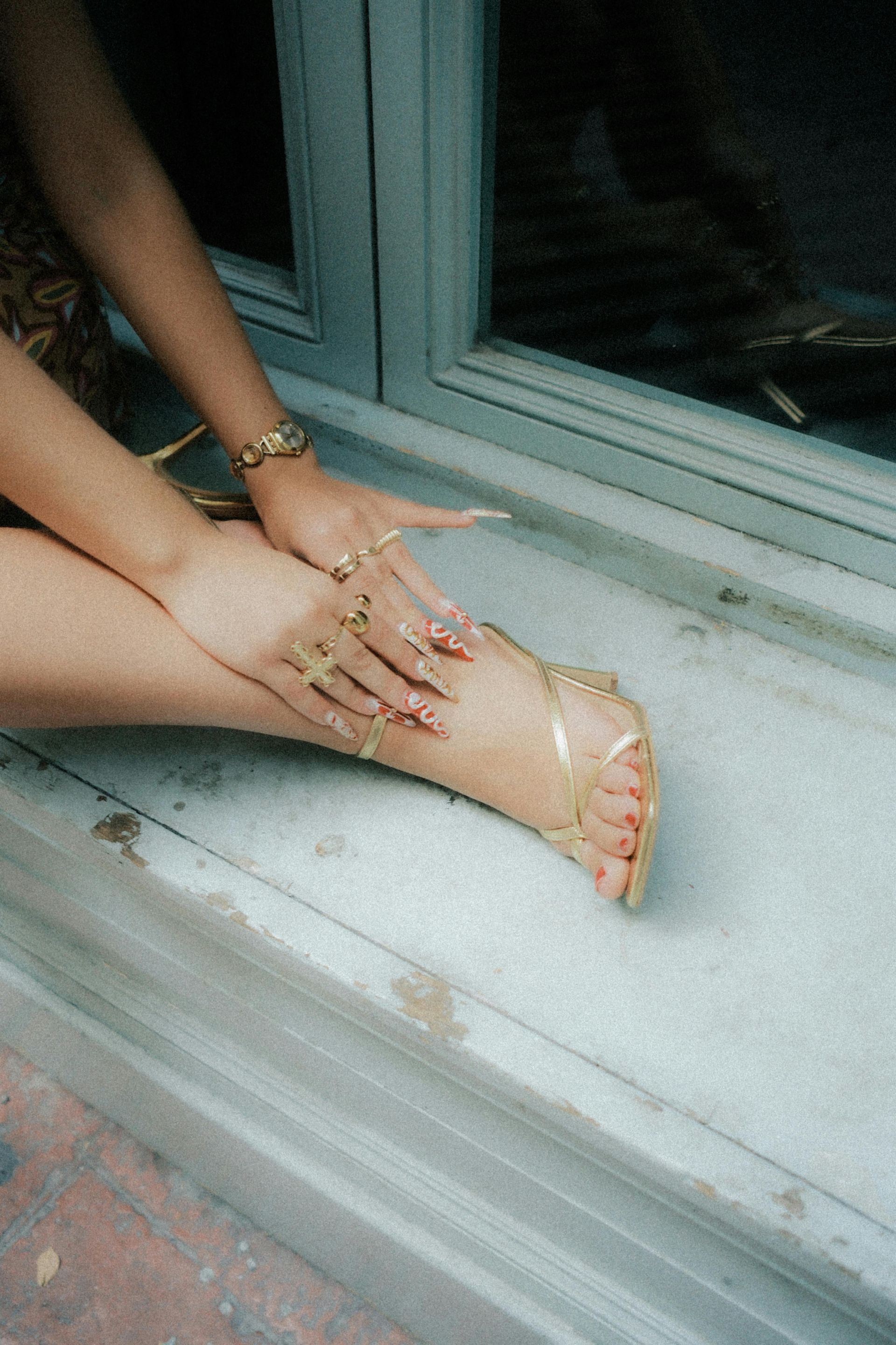 Dłonie i stopy osoby z długimi paznokciami, pierścieniami i złotymi sandałami spoczywają na parapecie okna.