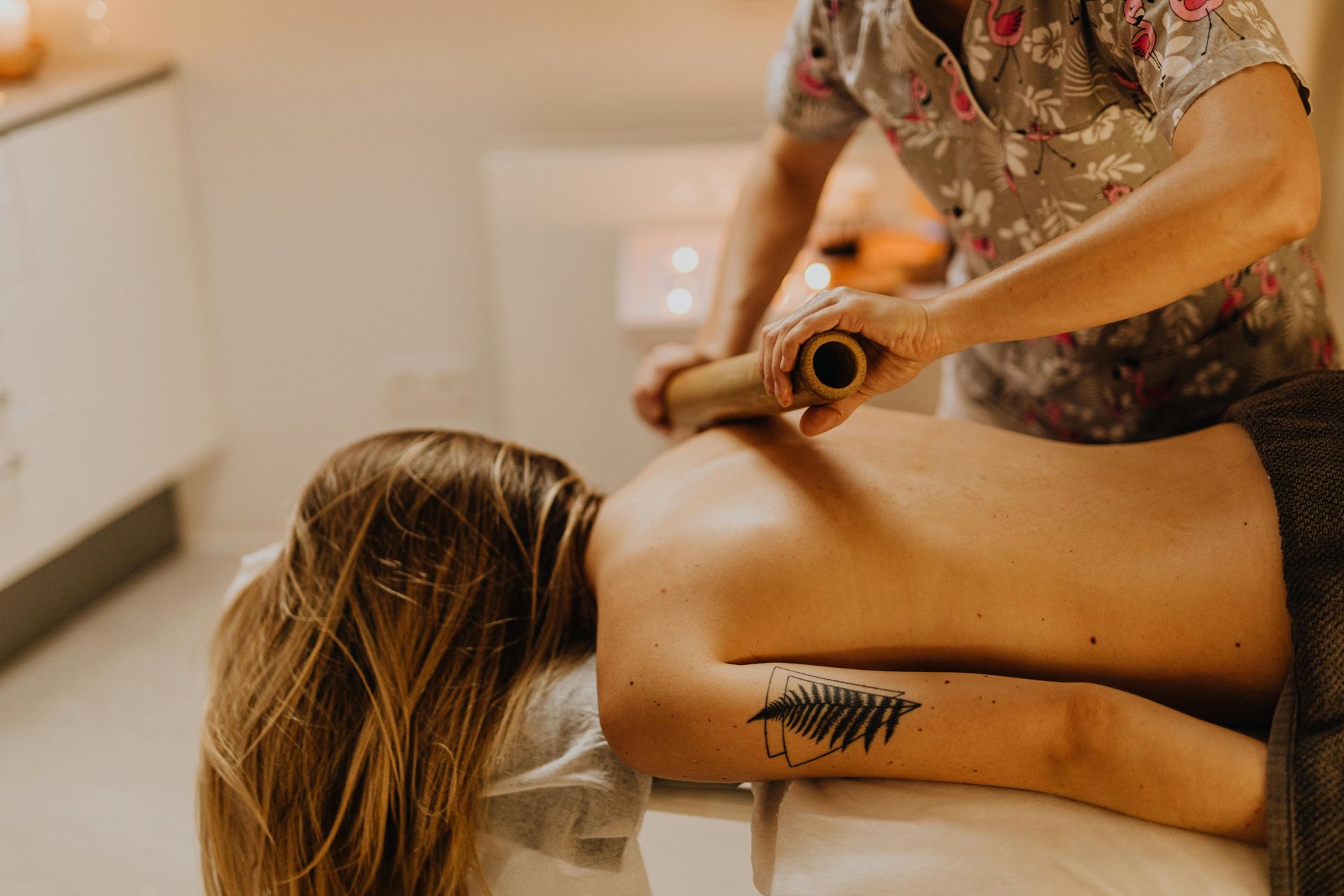 Osoba poddawana masażowi pleców bambusem w otoczeniu przypominającym spa, przy delikatnym oświetleniu.