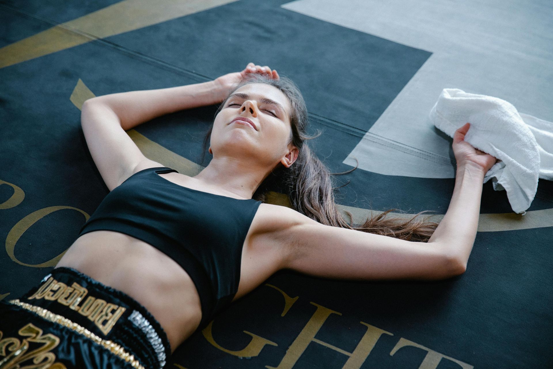 Woman in black sports bra and shorts rests on the floor, holding a towel, eyes closed.