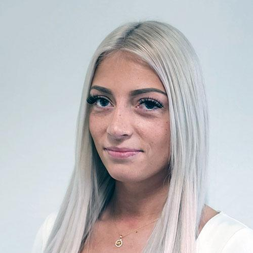 Porträt einer blonden Frau in einem weißen Oberteil vor einem einfarbigen hellen Hintergrund