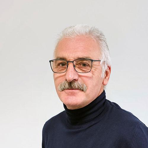 Porträt eines älteren Mannes mit weißem Haar, Brille und Schnurrbart, der einen dunklen Rollkragenpullover trägt.
