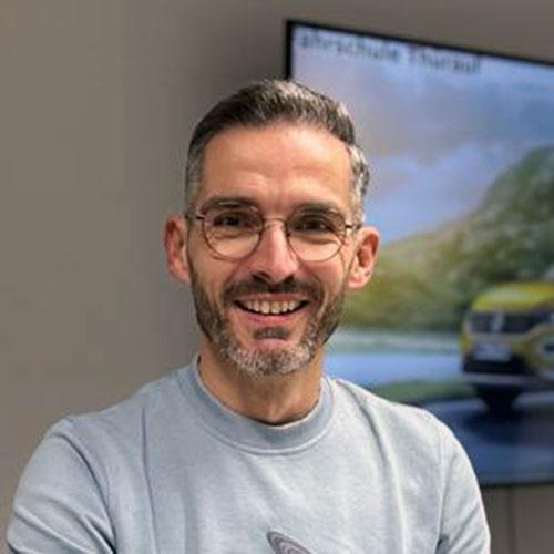 Ein lächelnder Mann mit Brille und grauem Sweatshirt in einem Büro, hinter ihm auf einem Bildschirm ein Autofoto.