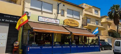 Cafetería española a pie de calle con terraza azul, toldo a rayas y banderas en un día soleado.