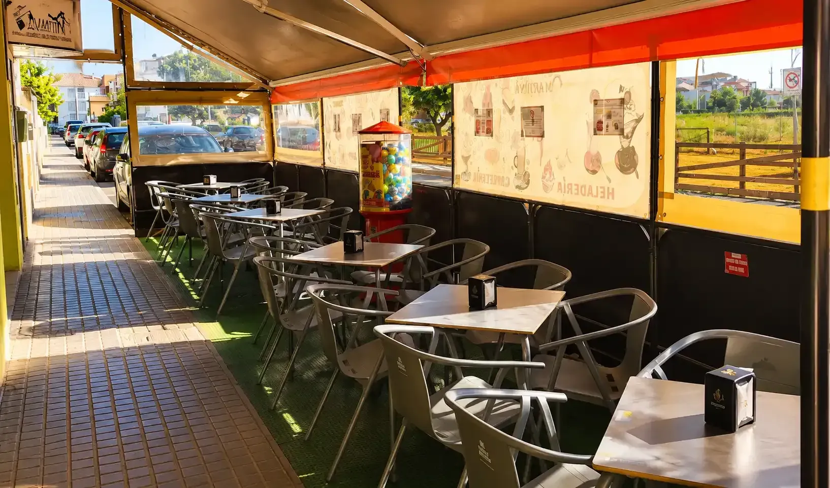 Terraza de cafetería con mesas y sillas bajo un toldo rojo junto a una acera y una carretera.