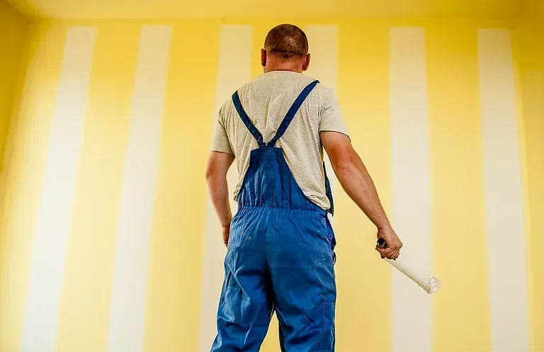 Un hombre está parado frente a una pared de rayas amarillas y blancas sosteniendo un rodillo de pintura.
