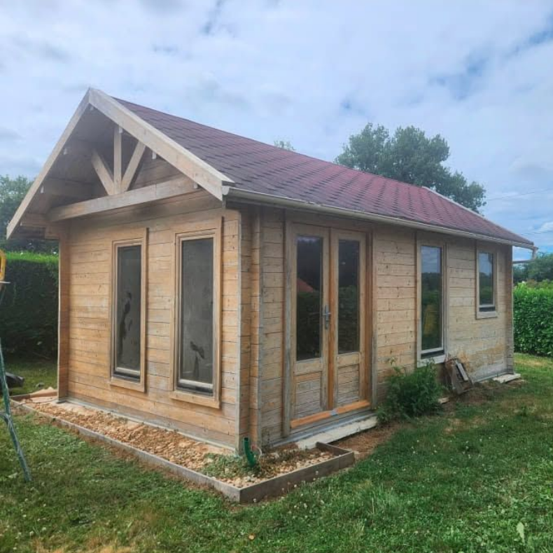 Petite cabane en bois avec un toit rouge et de grandes fenêtres dans une cour herbeuse