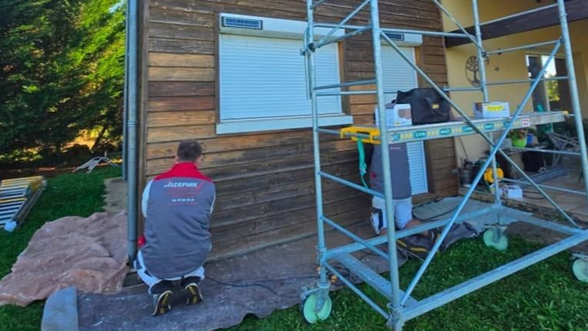 Une personne agenouillée près d'une maison à bardage en bois tandis qu'une autre travaille sur un échafaudage lors de réparations extérieures.