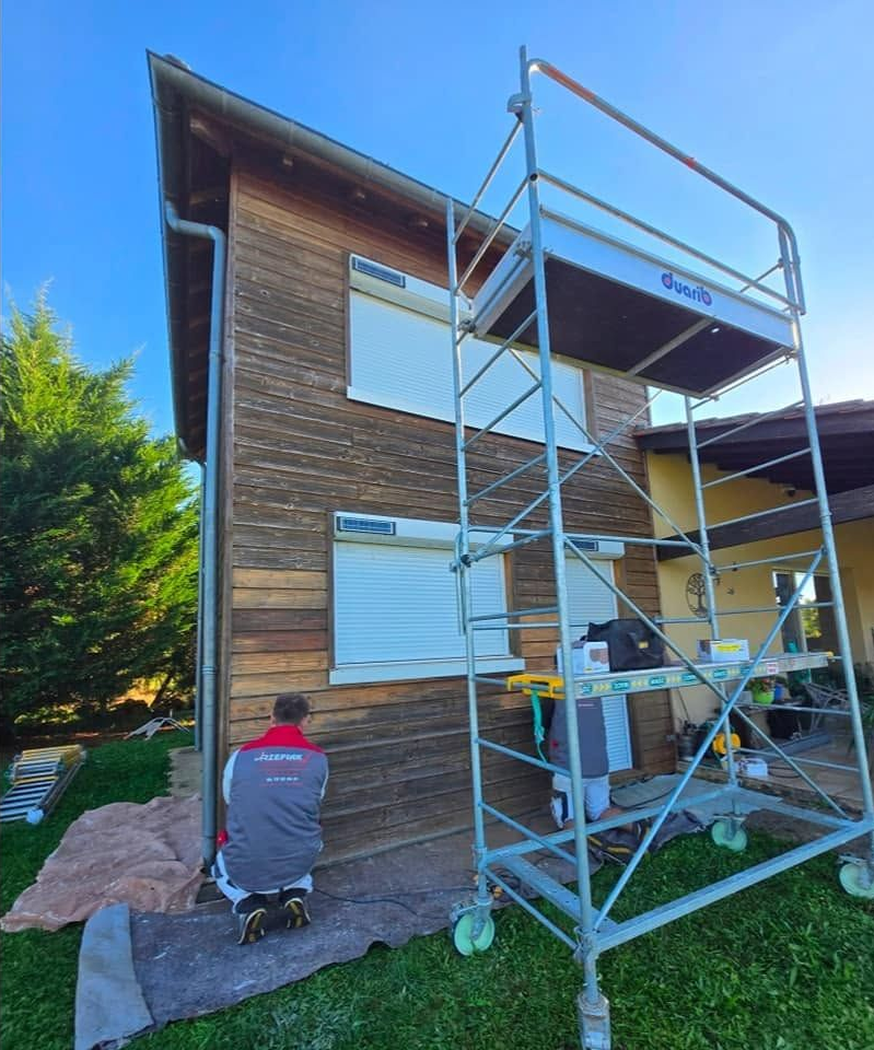 Une personne peint l'extérieur d'une maison en bois à côté d'un échafaudage par une journée ensoleillée