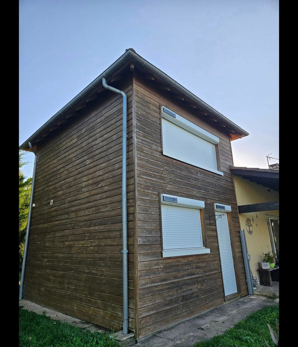 Petite maison en bois de deux étages avec volets fermés et descente de gouttière en métal, vue de côté depuis l'extérieur.