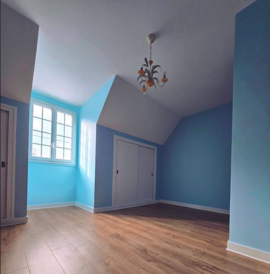 Chambre bleue vide avec parquet, plafond mansardé, fenêtre et lustre.