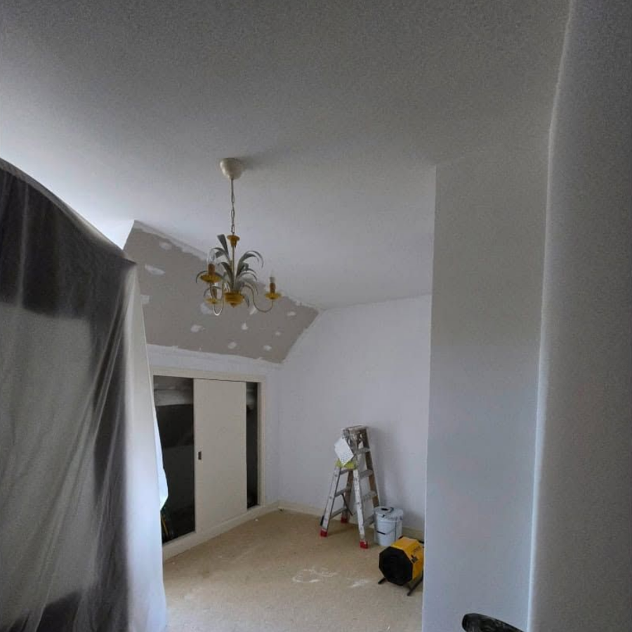 Pièce vide aux murs blancs, avec un lustre, une fenêtre occultée et une échelle sur laquelle se trouvent des fournitures de peinture.