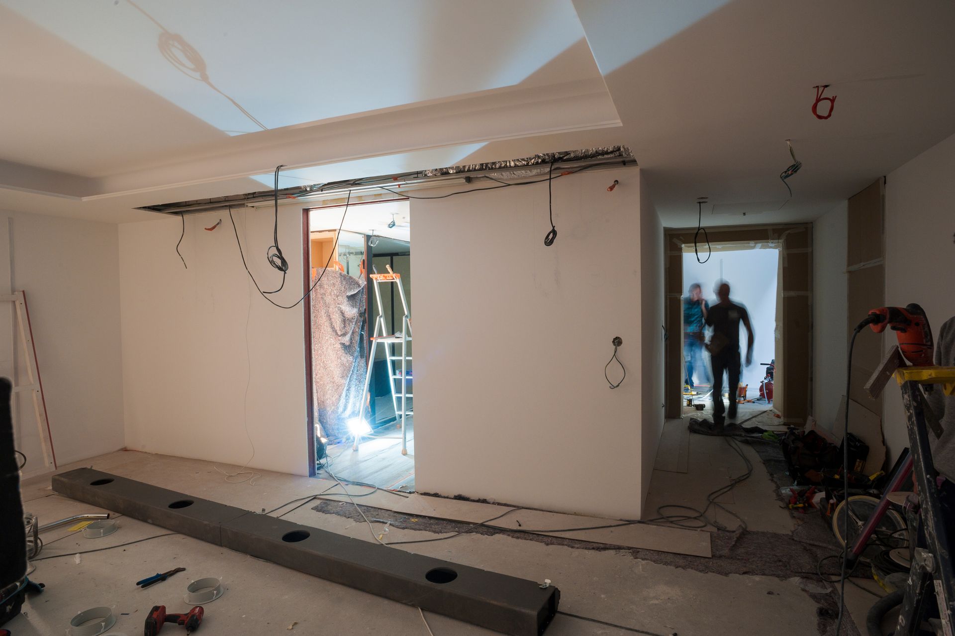 Chantier de rénovation avec murs inachevés, fils électriques apparents, échelles et ouvriers dans une porte et un hall