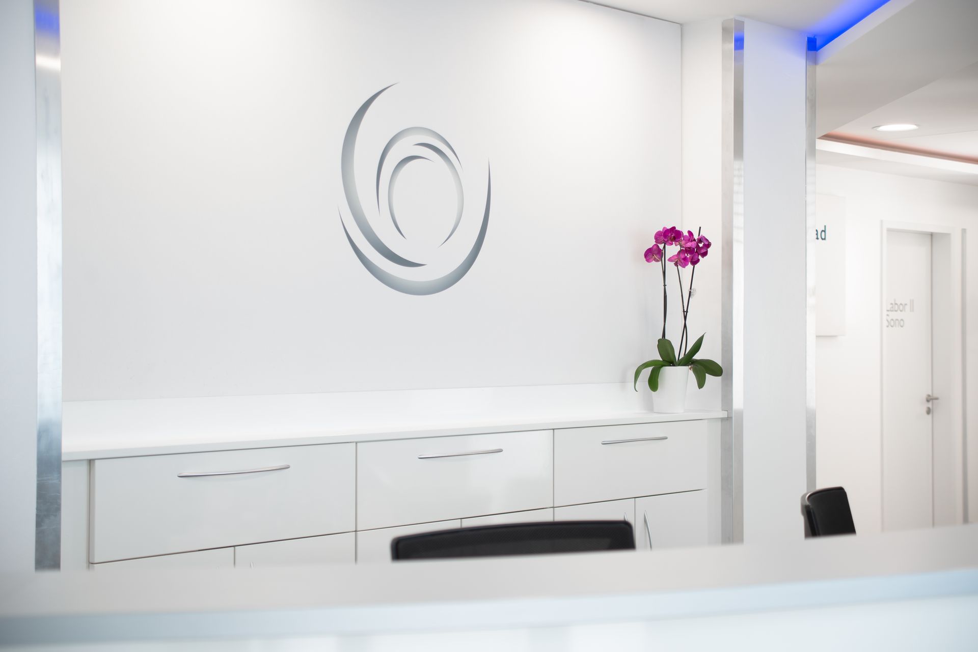 Ein Praxiszimmer mit Liege, Schreibtisch, Monitoren und einer Uhr an der Wand.