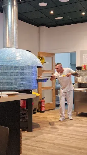 Un chef con uniforme blanco utiliza una pala larga para atender un horno de pizza, dentro de un restaurante.