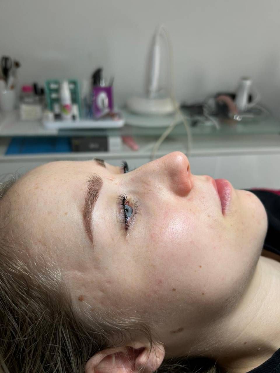 Femme aux yeux fermés, de profil. Teint clair, sourcils maquillés en brun, se reposant dans un salon de coiffure.