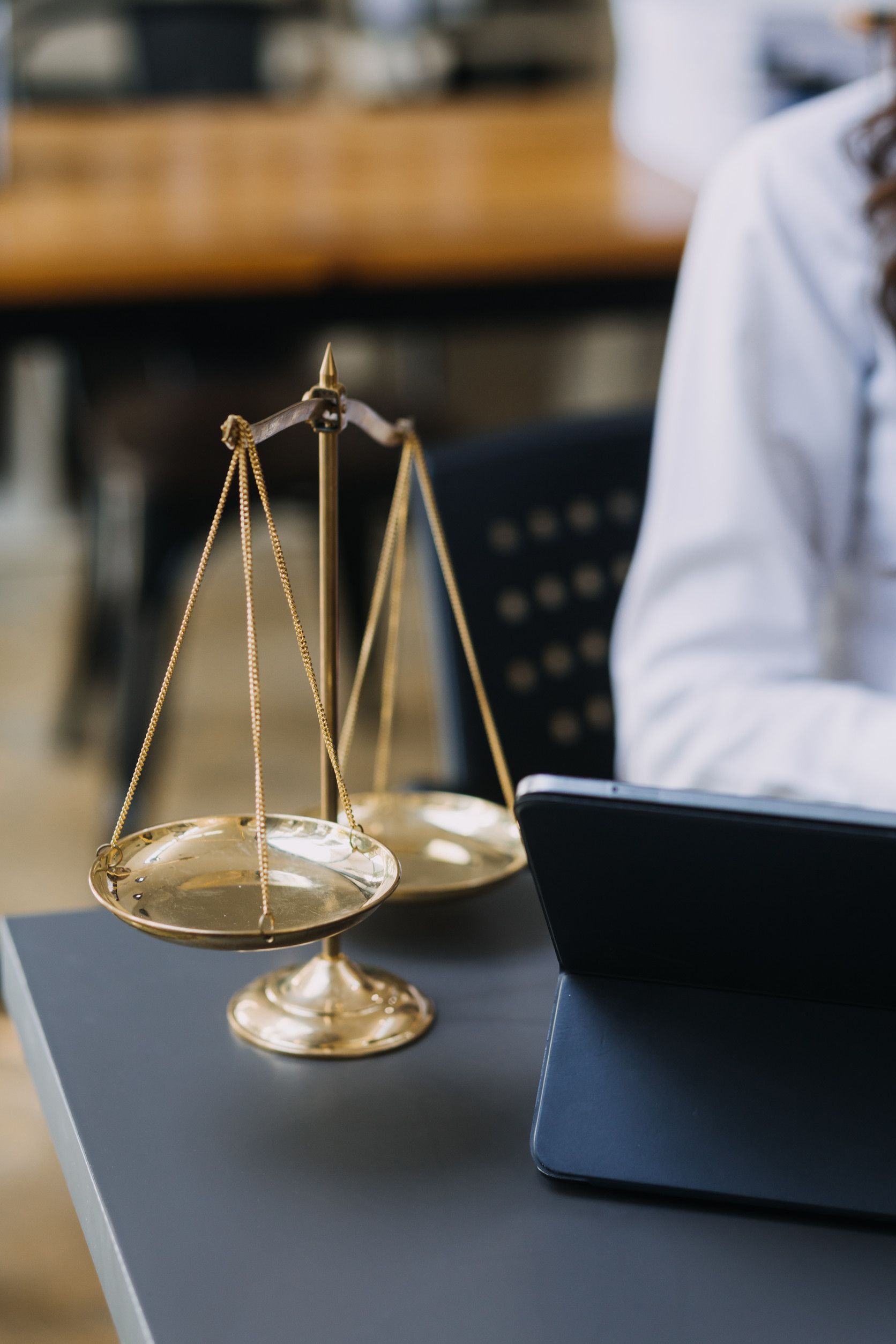 Balanza dorada de la justicia sobre un escritorio, con una persona y una tableta en el fondo.