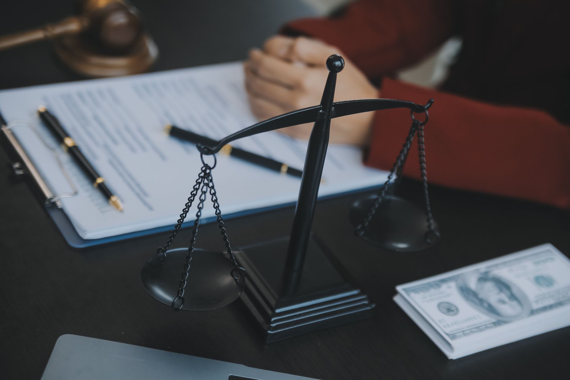 Una balanza de justicia negra sobre un escritorio con documentos legales, dinero y un mazo, con las manos de una persona.