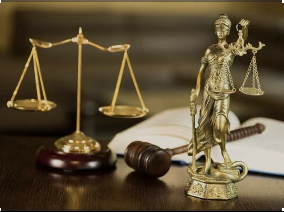 Una balanza de justicia negra sobre un escritorio con documentos legales, dinero y un mazo, con las manos de una persona.