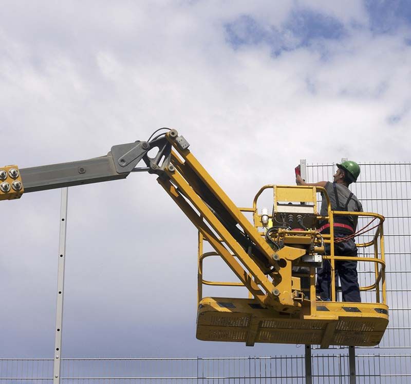Trabajador en una plataforma elevadora amarilla junto a una valla bajo un cielo nublado.