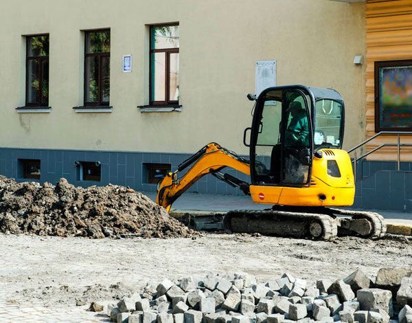 Miniexcavadora amarilla en una obra en construcción junto a un edificio, removiendo un montón de escombros.
