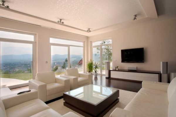 Luminoso salón moderno con sofás blancos, grandes ventanales, vistas a la montaña y televisor montado en la pared.