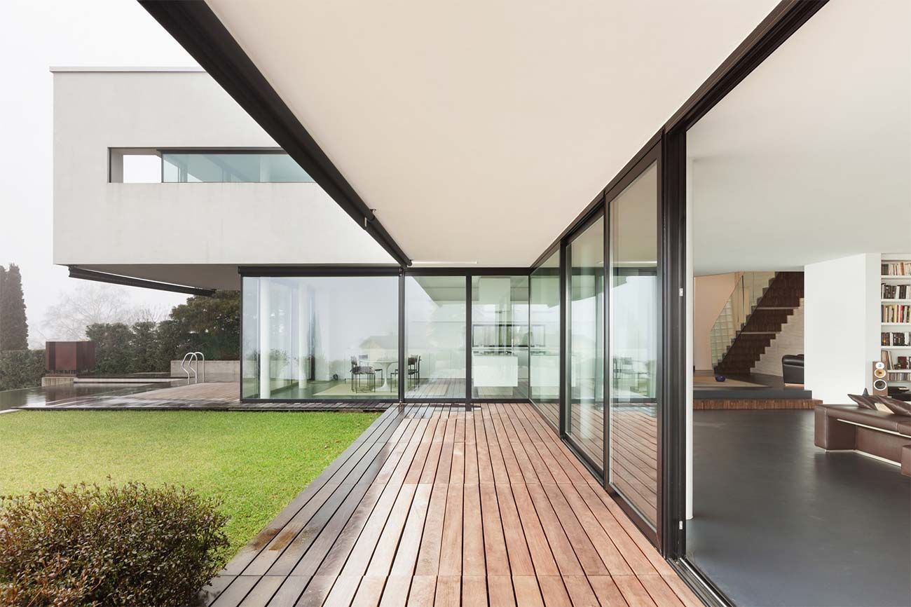 Interior de una casa moderna con paredes de cristal, terraza de madera y vistas al jardín exterior.