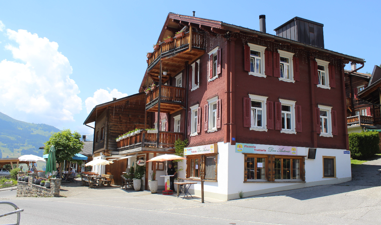 Ein rotes Holzhaus im alpinen Stil mit weiss umrandeten Fenstern an einer sonnigen Strasse mit Bergen im Hintergrund