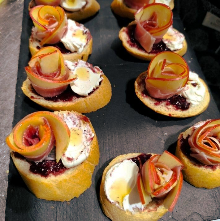 Aperitivos tostados con guarnición de rosa de manzana, queso de cabra y mermelada sobre una pizarra negra.