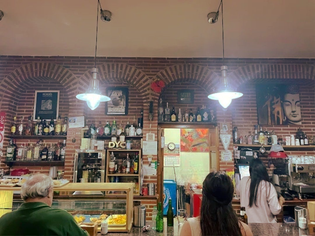 Interior de un café con una pared de ladrillos, botellas en los estantes y clientes cerca de un mostrador y una puerta.