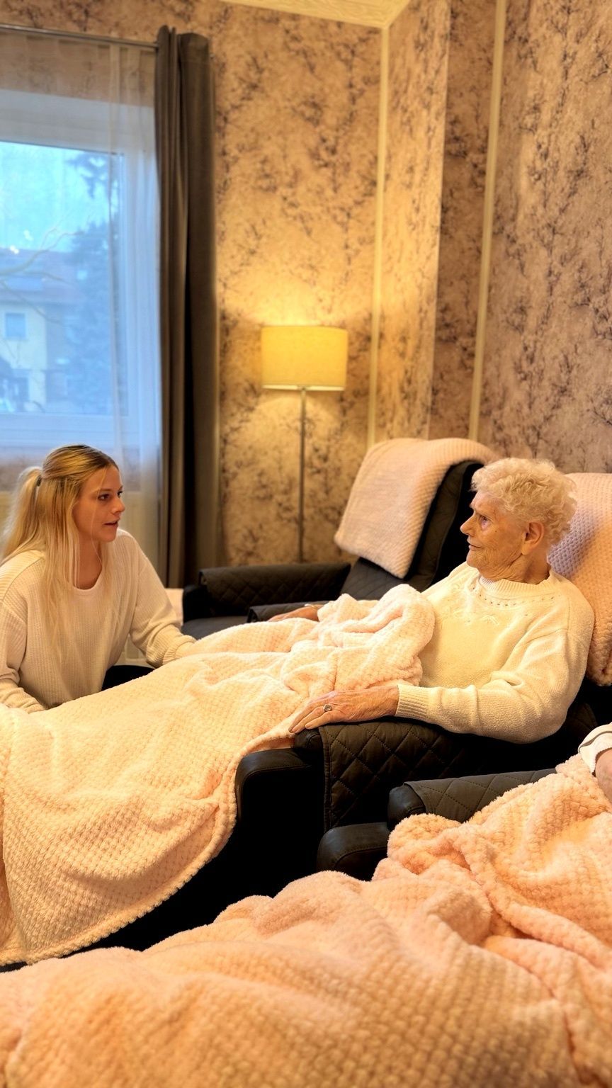 Eine Mitarbeiterin des Gesundheitswesens lächelt, während sie sanft die Schulter einer älteren Frau berührt, die auf einem Stuhl sitzt.