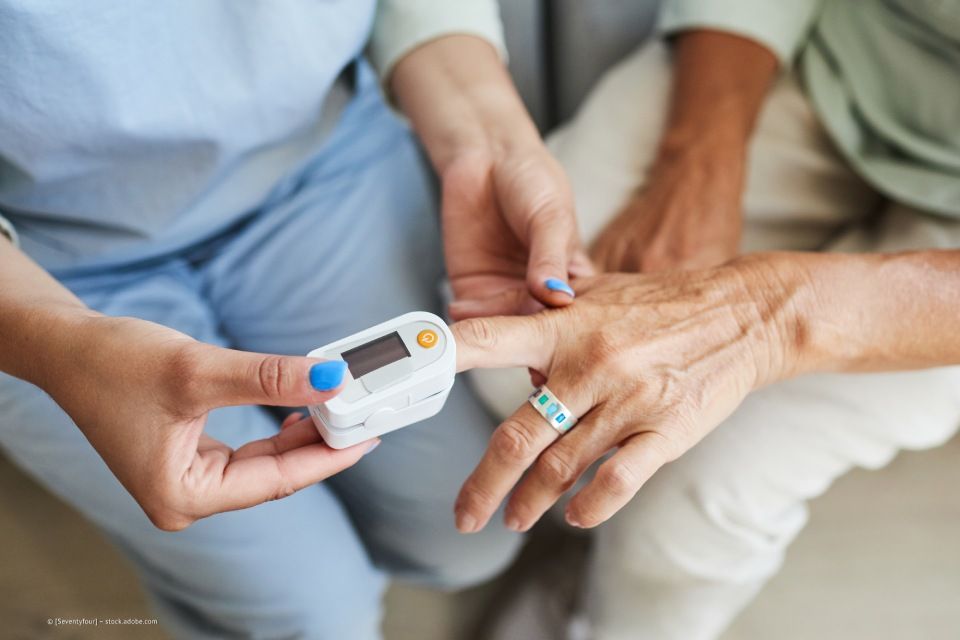 Eine Person benutzt ein Pulsoximeter am Finger einer anderen Person. Das Gerät zeigt ein Display an, und die Hände sind sichtbar.