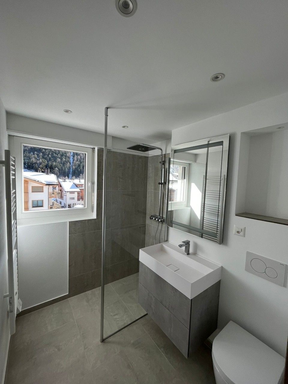 Bagno moderno con doccia, lavandino, specchio e finestra con vista sulle montagne innevate.