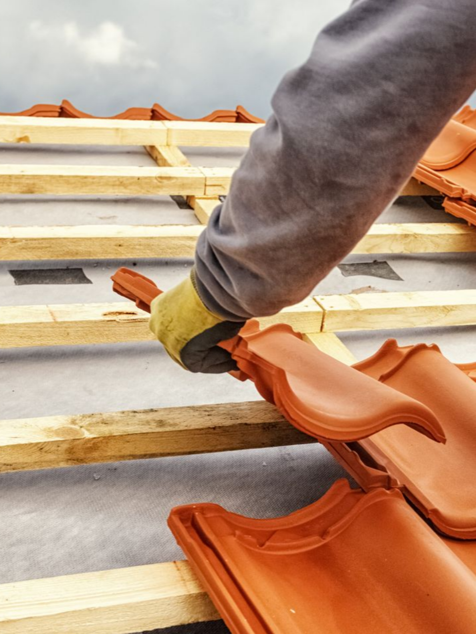 Une personne portant des gants de travail installe des tuiles de toit en terre cuite sur une charpente en bois.