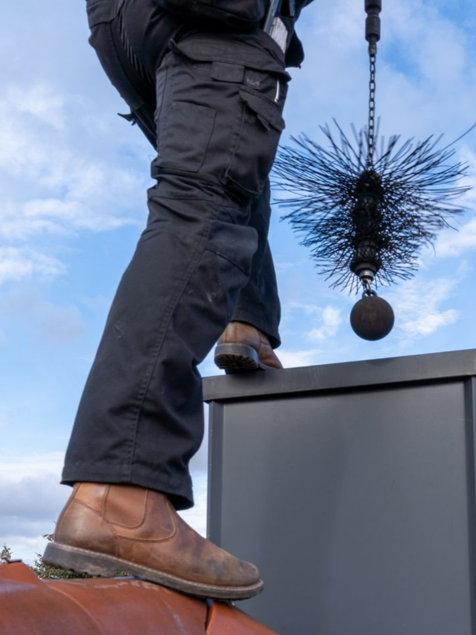 Personne sur un toit nettoyant une cheminée avec une brosse et une chaîne ; ciel nuageux en arrière-plan.