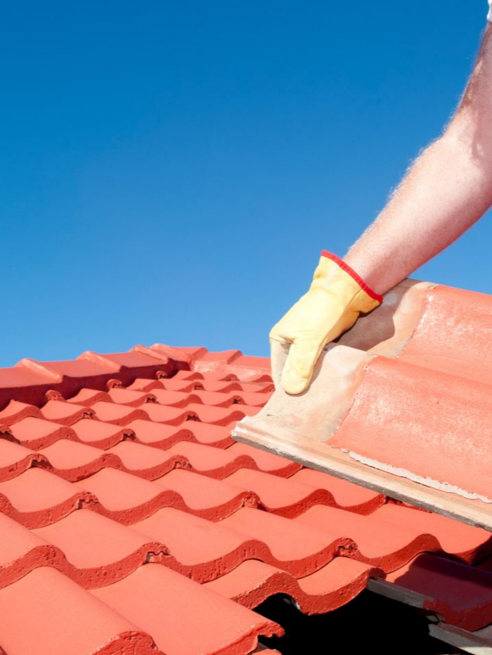 Une personne portant des gants pose des tuiles rouges sur un toit sous un ciel bleu.